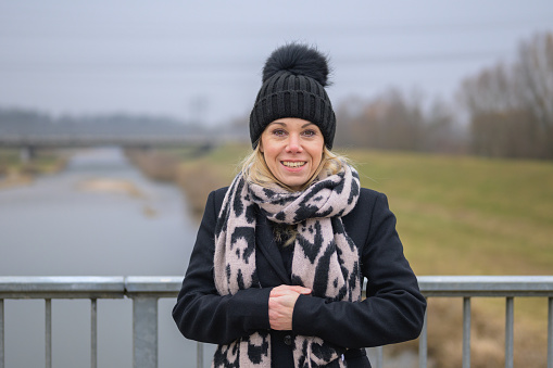 Attractive blonde lady wearing a black coat jacket and black bobble hat looks happily into the camera while standing on a bridge