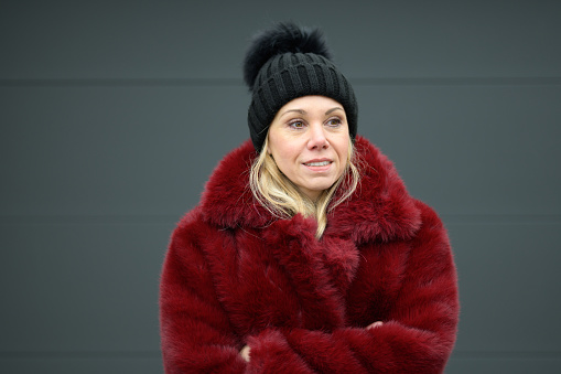 Attractive blonde lady wearing a red jacket and black bobble hat stands in front of a dark wall with her arms crossed