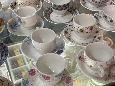 Assorted porcelain teacups and saucers with various patterns displayed on a glass table.