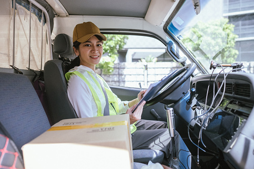 Happy Asian woman in casual attire, wearing a cap and a reflective vest, sits in the driver’s seat, checking a document pad to check information and prepare for a road trip to deliver packages and provide courier services. Looking at camera.