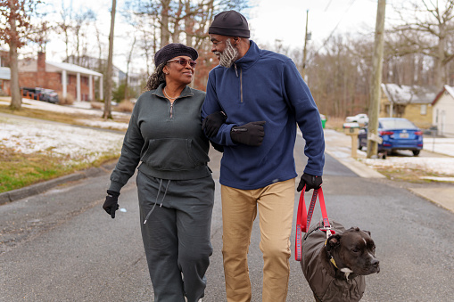 An active African American senior couple walk arm in arm and enjoy good conversation while taking their pet dog on a walk through their residential neighborhood on a cool winter day.