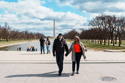 A vibrant senior couple walk hand in hand while touring Washington DC on a fun and relaxing winter vacation, with the Lincoln Memorial Reflecting Pool and Washington Monument visible in the background.