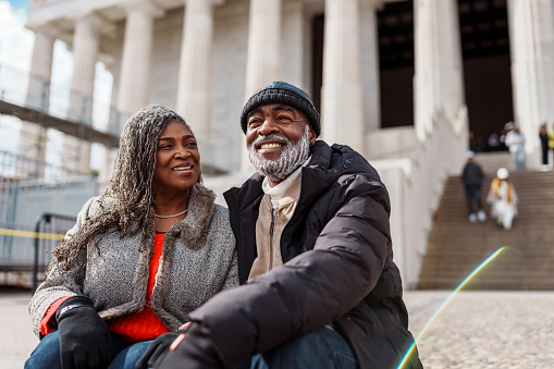A cheerful and well dressed senior couple of African American descent sit on the steps of the Lincoln Memorial while on a fun and relaxing winter trip to Washington DC.
