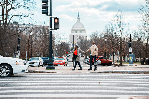 A vibrant senior couple of African American Descent hold hands and wait at a crosswalk while exploring Washington DC on a winter vacation, with a view of the United States Capitol visible in the background.
