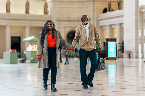 A senior couple of African American descent smile and hold hands while walking through a train station with a small piece of luggage during a weekend getaway to Washington DC.