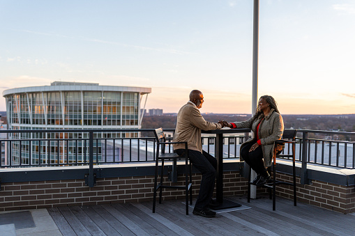 An affectionate and vibrant senior couple of African American descent sit at a table on the rooftop of their luxury hotel in Washington DC and enjoy good conversation with sunset views over the city.