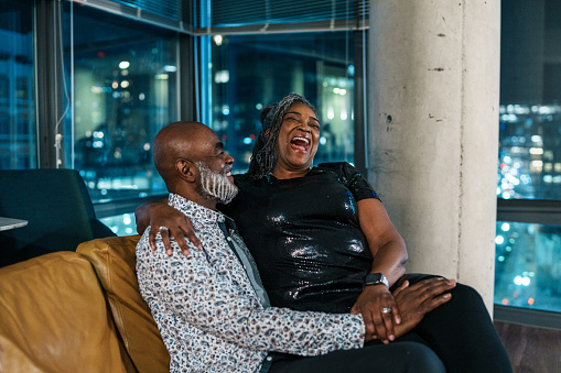 A well-dressed senior couple of African American descent affectionately snuggle on the couch in their luxury hotel room and laugh after spending en evening out on the town during a weekend getaway to Washington DC.