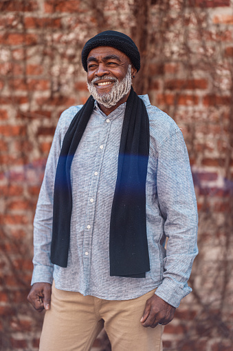 Portrait of a vibrant and handsome African American senior man wearing a hat and scarf,  smiling while standing in front of a brick wall outside.