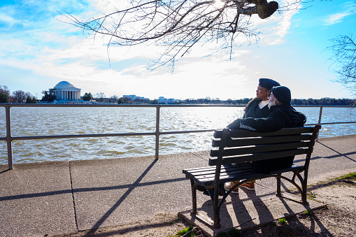 An active and loving senior couple of African American descent sit on a park bench and enjoy the scenic view of the Jefferson Memorial while on a relaxing walk in Washington DC in the winter.