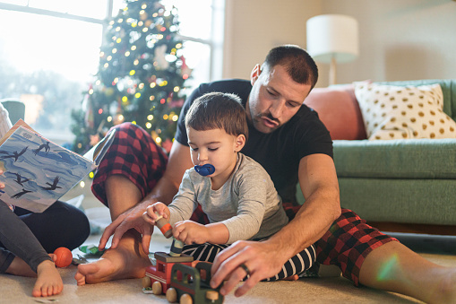A playful and affectionate young dad and his multiracial toddler son of Puerto Rican descent play with a wooden toy train together on a cozy and lazy winter day at home, with a tree decorated for Christmas visible in the background.