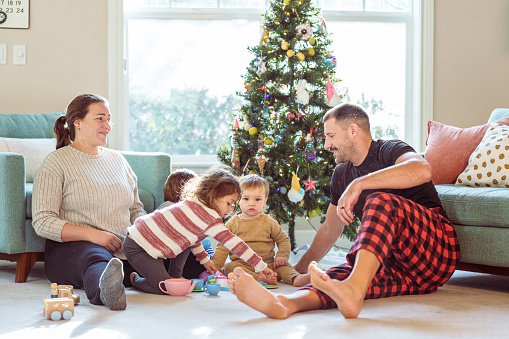 A Puerto Rican woman and her husband spend Christmas at home with their three young children who are playing with a toy tea set and wooden cars in front of the Christmas tree.