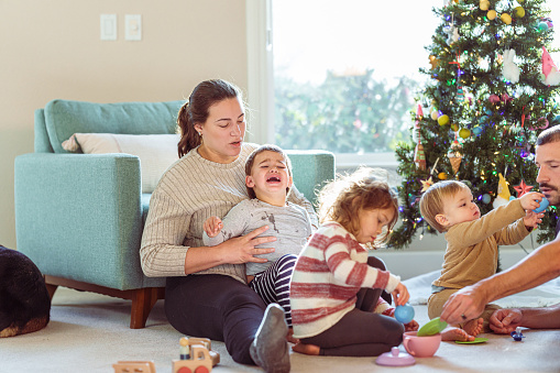 A Puerto Rican woman affectionately holds and consoles her crying two year old son as her husband plays with their other two children. The family is celebrating Christmas together at home in their living room.