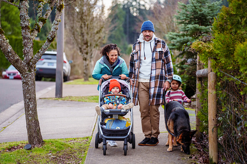 A cheerful Eurasian woman pushes her toddler son in a stroller while on a relaxing walk with her husband, four year old daughter and German Shepard pet dog along a sidewalk in their residential neighborhood located in Oregon on a cool, winter day.