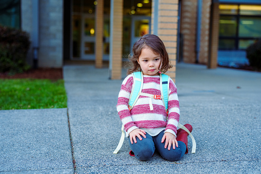 A cute four year old Eurasian girl wearing a backpack sits on the sidewalk in front of her school and patiently waits for the doors to be unlocked and school to begin.