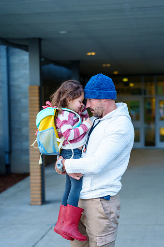 A handsome young dad, who walked his Eurasian preschool age daughter to school, gives her a hug goodbye before she heads into the school building.
