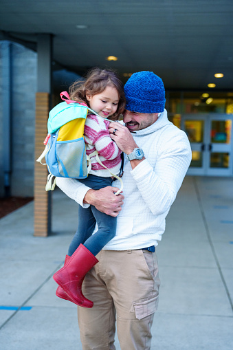 A handsome young dad, who walked his Eurasian preschool age daughter to school, gives her a hug goodbye before she heads into the school building.