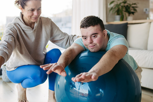 Young man with Down syndrome exercising at home with his mother on a fitness ball. Workout routine for disabled man.