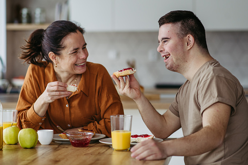 Young man with Down syndrome having breakfast with his mother at home. Morning routine for man with Down syndrome genetic disorder.