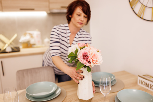 Middle aged woman putting on vase with flowers while setting the table for dinner at home