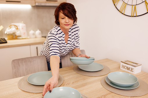 Middle aged woman putting on plates while setting the table for dinner at home