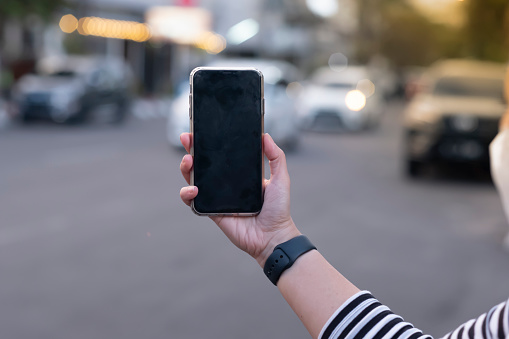 Horizontal image of female hand holding a smartphone with blank screen. Oily of black smart phone. Evening city street on the background, bokeh light.