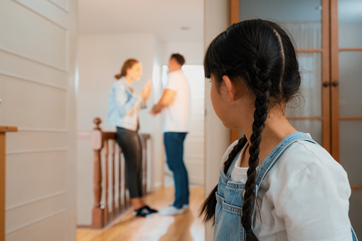 Stressed and unhappy young girl huddle in corner crying and sad while her parent arguing in background. Domestic violence at home and traumatic childhood develop to depression. Synchronos