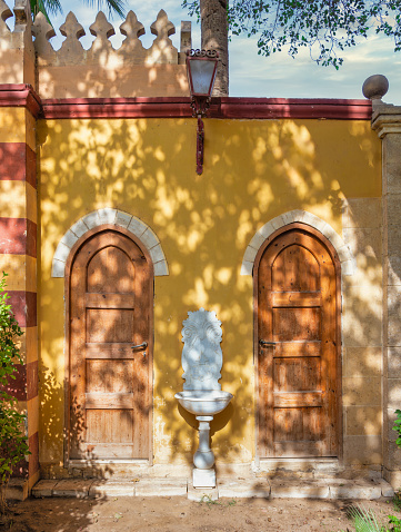 Prince Naguib Palace: Pair of weathered wooden doors stand nestled against a vibrant yellow wall. A gently trickling fountain mediating the doors adds a touch of tranquility to the scene
