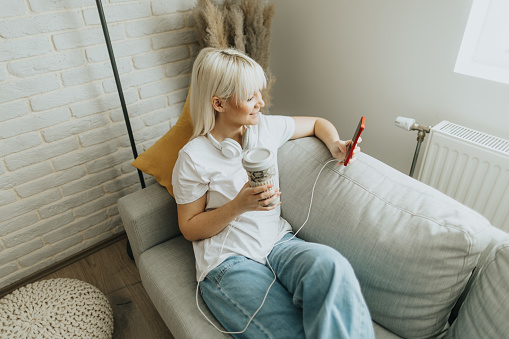 High angle view of beautiful young blond woman drinking coffee and using smart phone while relaxing on sofa at home