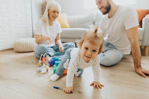 Portrait of cute baby girl looking at camera while crawling at home while smiling young parents sitting on floor in background