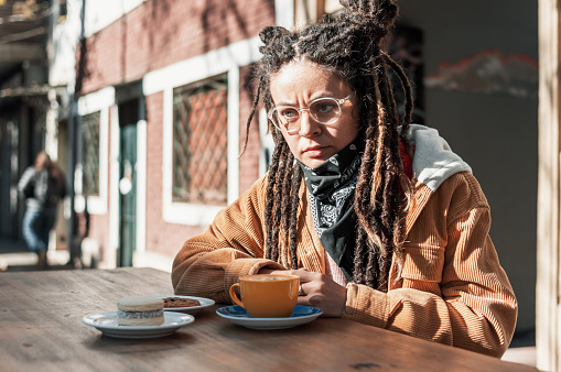 young latina woman with dreadlocks of colombian ethnicity, wearing a brown jacket and glasses, is sitting outside the cafeteria drinking coffee.