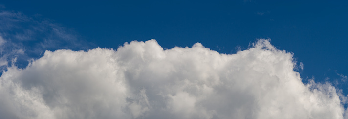 Fat cumulus clouds accumulate to build an enormous mountain of clouds, announcing heavy weather. A sky only, full-frame and close-up image of a tremendous cumulonimbus cloud. Some cirrus clouds appear high up in the blue sky and a band or roll cloud of stratocumuli extend low over the foreground. A sky only, full-frame and close-up image.