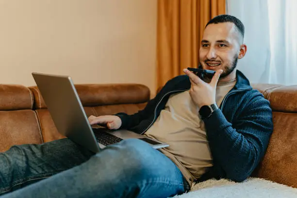 From his home office, a man adopts a dual-screen approach to productivity, his focus divided between the laptop and mobile phone, maximizing efficiency and connectivity in his professional endeavors