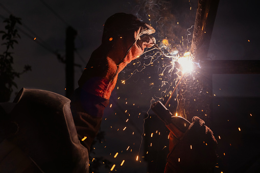 A man is entertaining a crowd by welding a piece of metal with a bright lens flare in the dark, creating a spectacle of heat and music at midnight