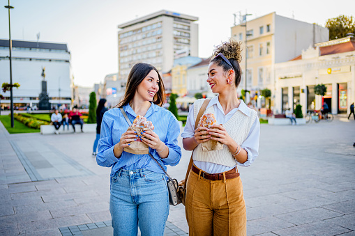 Young cheerful female friends walking and eating pastries in the city