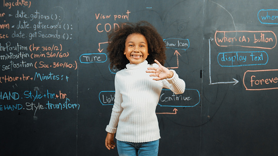 Happy african girl writing blackboard and turnaround to waving at camera while standing at board with engineering prompt or coding, programing system written in STEM technology classroom. Erudition.