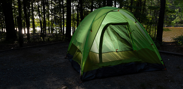 Campsite in the trees with plenty of shade and a lighted tent with a lake in full sunlight behind.