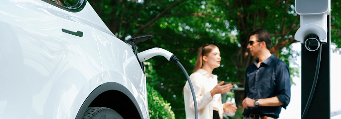 Young couple recharge electric car battery from charging station in green city park in springtime. Rechargeable EV car for sustainable environmental friendly urban travel lifestyle. Panorama Expedient