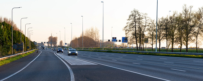 Netherlands, South Holland, Reeuwijk, April 19th 2023, Dutch gray 2022 electric Tesla Model 3 and other traffic (cars, trucks, vans) driving on the A12 multiple lane highway between Gouda and Reeuwijk at sunset on a sunny day in springtime, the A-12 is a 137 kilometer long highway crossing the country from The Hague in the East to Germany in the West