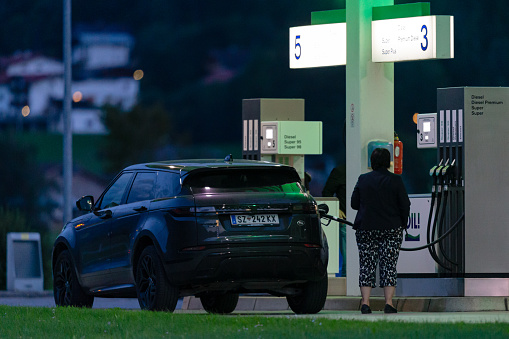 Austria, Tyrol, Zillertal, Uderns, August 8th 2023, nighttime side/rear view close-up of a senior woman refuelling an Austrian black Land Rover Range Rover SUV (from the Schwaz district) with Diesel fuel at the 