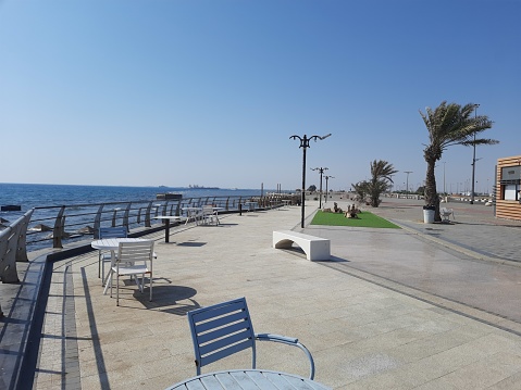 A corniche with tables, chairs and palm trees on a rocky shore of a calm sea and a clear blue sky on a sunny afternoon on the Red Sea shore