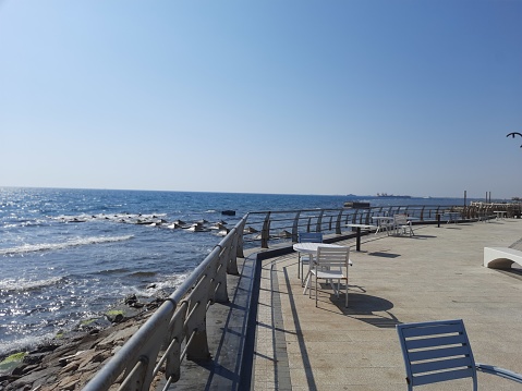 A corniche with tables and chairs on a rocky shore of a calm sea and a clear blue sky on a sunny day in the afternoon on the shore of the Red Sea