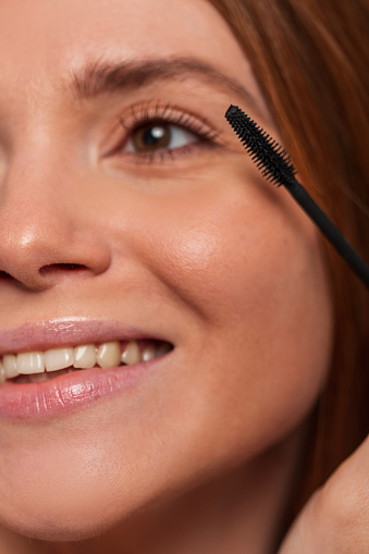 Beautiful happy redheaded woman model with a cute smile paints her lashes with mascara, closeup. Beauty and makeup