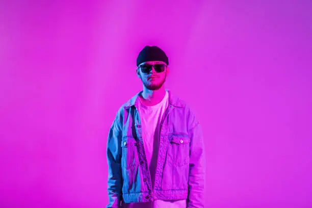 Photo of Stylish handsome creative young guy with a cap and sunglasses in a trendy fashion denim jacket in a studio with pink and neon lights