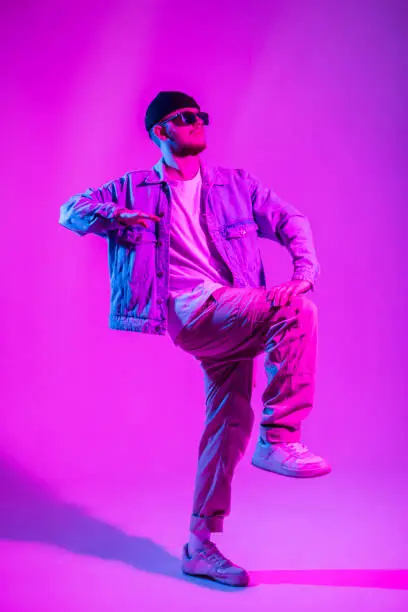 Photo of Fashionable handsome young dancer hipster hip hop guy with sunglasses in trendy fashion casual clothes with jeans jacket, pants and sneakers dancing in a color studio with creative pink and blue light