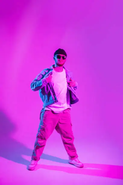 Photo of Stylish handsome young male dancer with fashion sunglasses and hat in trendy denim clothes is dancing in colored studio with pink and blue light