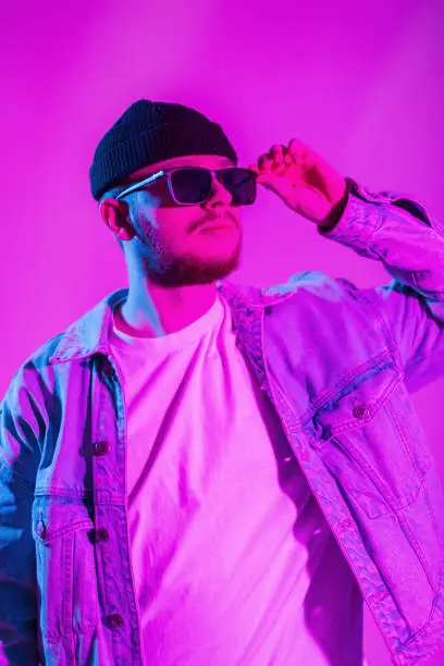 Photo of Stylish trendy handsome dancer man in trendy clothes with a fashion denim jacket and a T-shirt with a cap puts on stylish sunglasses on a pink background with creative colorful blue light.