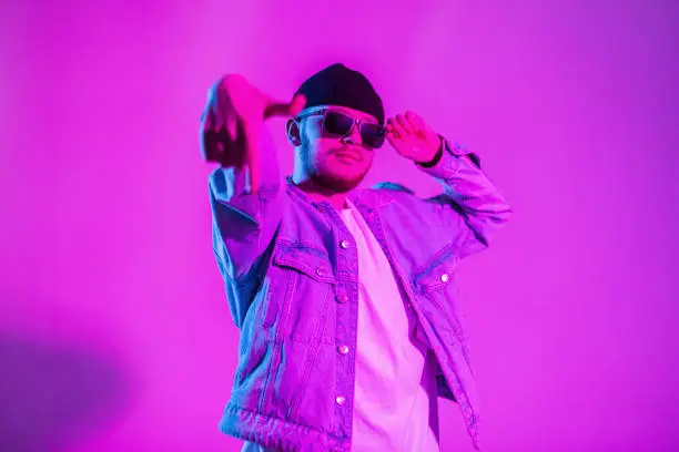 Photo of Cool dancer stylish man in trendy fashion denim clothes dancing and wears sunglasses in creative colored studio with pink and neon light