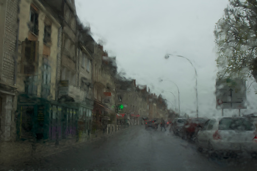A picturesque scene capturing a blurry road through a wet windshield on a rainy day in a charming french village. Parked cars line the right side, while medieval-style buildings adorn the left.