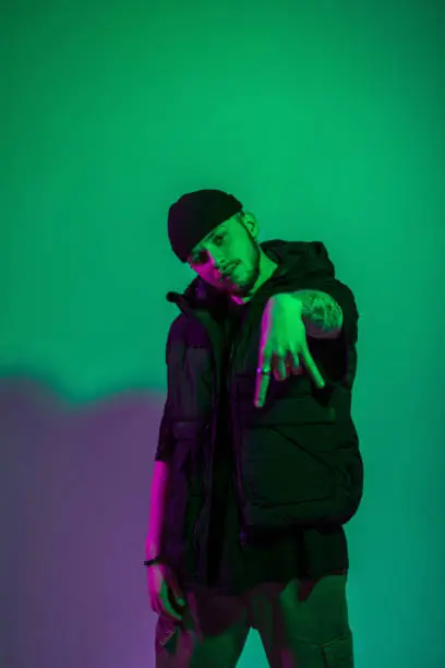 Photo of Fashionable handsome stylish man model dancer with a hat in a vest and black clothes with creative pink and purple light