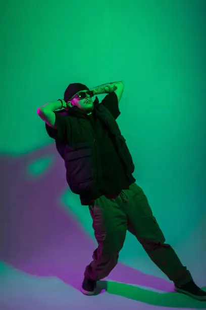 Photo of Young handsome stylish professional dancer guy with a stylish hat and sunglasses in fashionable black clothes dances and poses in a creative studio with green and pink lights